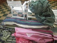 Photo showing white plastic patio chairs stacked and striped chaise lounge cushion with outdoor fabric items and umbrella parts on a checkered floor.