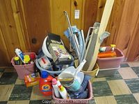 Wide shot of two plastic bins filled with assorted cleaning products and tools including a dustpan, mop handle, and bottles.