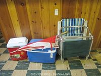 Red and blue coolers, two lawn chairs, and a folded red and white umbrella arranged against wooden paneled wall on checkered floor.