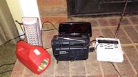 Photo showing red emergency flashlight, tall white emergency LED light plugged in, black hand-crank solar Observer radio flashlight, black Westclox digital alarm clock radio