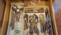 Drawer of assorted silverware pieces with ornate handle designs, identifiable as Oneida, alongside other flatware.