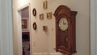 View of the wooden Howard Miller wall clock with arched top, six small framed pictures, and two wooden octagonal thermometers hung on the wall adjacent to a doorway.