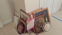 Photo of a collection of framed pictures leaning against the wall, visible various sizes and subjects including owls and floral with wood and metal frames.