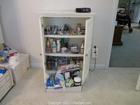 White wicker-front storage cabinet with both doors open showing shelves filled with personal care products, and some items outside the cabinet on the floor.