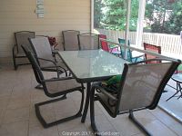 Patio table on covered porch with six beige mesh sling chairs visible around it, table has a frosted textured glass top with a dark metal frame and cantilever style chairs around it.
