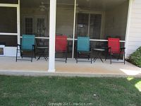 Four folding patio chairs (two red, two blue-green) and two small round frosted glass folding tables arranged on a porch.