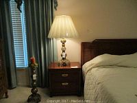 Full view of handcrafted brass Moroccan lamp standing on a bedside table next to a bed and candle holder.