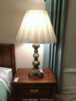 Full view of the brass Moroccan lamp on a wooden side table with the lamp turned on, showing the new white pleated lampshade and the intricate brass base.