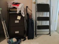 Brown 2-drawer file cabinet beside step ladder, black night stick, and suitcase in corner of room