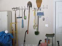Room corner showing yellow hand truck, cleaning tools hung on a wall rack including shovel, broom, rake, duster, dustpan set, and cleaning mitt.
