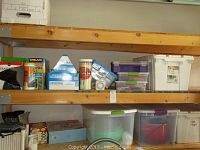 Shelf with various boxes and household items including Kirkland and Puffs boxes, trash bags, plastic containers with tools and miscellaneous items