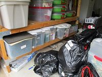 View of shelving unit with multiple plastic storage bins and black bags containing Christmas decorations and storage boxes.