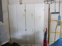 Two white double-door storage cabinets standing side by side in a room with tools and equipment nearby.