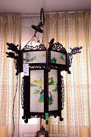 Full view of the Asian style hanging light suspended in front of sheer curtains showing the carved wood frame and scenic painted glass panels.