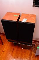 Two Mission Electronics 737 speakers with wood veneer tops and black fabric grills standing on worn black stands on wooden floor.