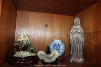 Shelf overview showing all four items: terracotta robed figure, children on buffalo figurine, porcelain jar on wood stand, stone pillow