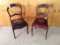 Two wooden chairs with floral needlepoint seats and carved wooden backs, placed on carpet against wooden wall.