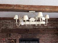 View of white metal farm motif light fixture installed on ceiling with six white lampshades and metal animal silhouettes, mounted over exposed wood beams and brick wall.