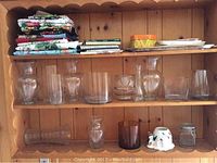 Wooden shelf holding multiple glass vases of varying shapes and sizes, layered tablecloths, and cookbooks on top shelf.