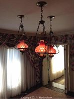 Three hanging hurricane style lights with rose-colored hobnail glass globes suspended by brass chain fixtures from the ceiling in a room with floral wallpaper and curtains.