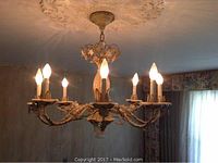 Gold and ivory metal chandelier with 8 lit candle-like lights, hanging from a ceiling medallion with floral design. Shows detailed leaf and floral metalwork.