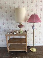 Photo showing two lamps and a small rolling table with a glass top decorated with seashells, against floral wallpaper.