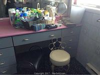 Photo of pink bathroom counter with multiple cleaning liquid bottles, spray bottles, soap dishes, toilet paper roll holder, and a decorative metal waste basket beside the counter.