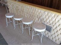 Four white chairs lined up against a wall, all with circular wooden seats and metal backs with circular design.