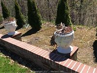 Pair of white cement urns placed on brick pillars in a garden. Both urns show wear and cracks, contain dried foliage and are decorated with a Greek key pattern.