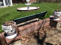 Two cement urns placed outdoors on brick pillars with lawn and fountain visible in background. One urn is closer and 16 inches in diameter, the other further and 19 inches tall. Both show wear and hold dry plant debris.