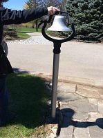 Metal bell mounted on a post, showing the full height and installation into stone paving.