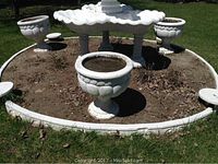 Overview photo of the three white cement garden urns placed in a garden bed surrounding a large white fountain or planter.