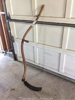 Full view of the antique scythe leaning against a garage door, showing curved wooden handle and metal blade.