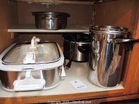 View inside cabinet showing two crockpots (one rectangular casserole style and one standard round) and other stainless steel cookware items.