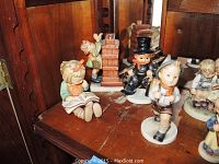 Overall view of nine Hummel figurines on wooden surface showing sitting girl reading, chimney sweep boy, boy walking and group reading