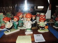 Six Charlot by Goebel figurines with red hair, various poses including doctor, graduation, and children playing. Photographed on wooden surface with mirror background.