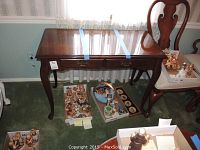 Front view of the wooden side table showing polished surface, drawer, and curved legs. Plates of small collectible figurines sit underneath the table as seen in the background.