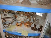 Photo showing assorted gold plated tea set pieces, salt and pepper shakers, and casserole dishes on white shelf.
