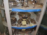 Full view of silver serving ware in cabinet showing covered serving bowl on stand, trays, bowls, and smaller condiment dishes.