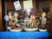 Front view of four Goebel Hummel figurines inside a glass cabinet showing their front features and colors