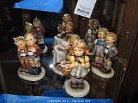 Four Goebel figurines displayed inside a wooden cabinet with glass shelves reflecting behind them