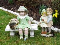 Overview showing both lawn statues placed on grass near flower beds and white stone border. One is of a girl on a bench, another of two children together.