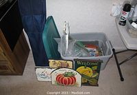 Plastic bin containing several painted decorative slate signs propped against it, including Home Sweet Home, Happy Fall pumpkin, and Welcome with bananas signs.