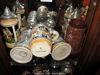 View of six beer steins displayed on a glass shelf inside a wooden cabinet with mirrored back. Includes five colorful ceramic steins with detailed painted scenes and one brown relief embossed ceramic stein. Pewter lids with thumb levers visible.