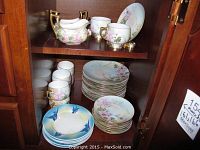 Porcelain dinnerware set including floral cups, creamer, and plates stacked on shelves inside a cabinet