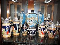 Full group of six German beer steins displayed on a glass shelf inside a cabinet, showing variety in designs, colors and lid styles.