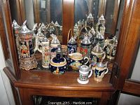 Full view of 9 German beer steins and sugar and creamer set on wood cabinet shelf with mirror behind