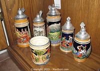 Set of five German beer steins and one 2-handled mug displayed on wooden shelf, showing front painted scenes and pewter lids