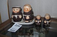 Front view of four monk-themed ceramic pieces: creamer, sugar bowl on tray, salt shaker, and pepper shaker.