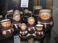 Frontal view of six monk figurines arranged showing size and details, with faces and hands painted on ceramic material in shades of brown and cream.